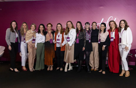 Fotografia de doze mulheres fundadoras e diretoas da Weem em pé, lado a lado, posando para a câmera. Todas usam crachás do Weem Talks pendurados no pescoço e vestem roupas formais de cores variadas, como branco, preto, vermelho e bege. Elas sorriem, em frente a uma parede de fundo roxo no local do Weem Talks dois mil e vinte e quatro.