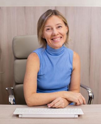 Mulher sorridente com blusa azul sem mangas, sentada à mesa com teclado branco, em ambiente claro.