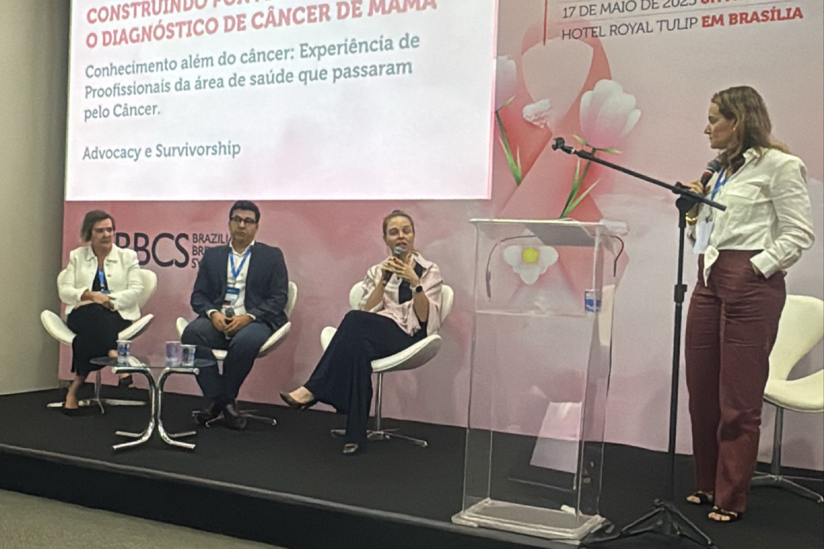 Painel com quatro profissionais debatendo experiência pós câncer de mama diante de banner rosa.