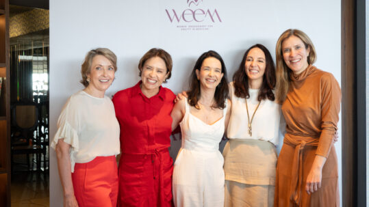 Cinco mulheres sorridentes abraçadas diante do logo WEEM, vestindo tons claros, vermelho e marrom.