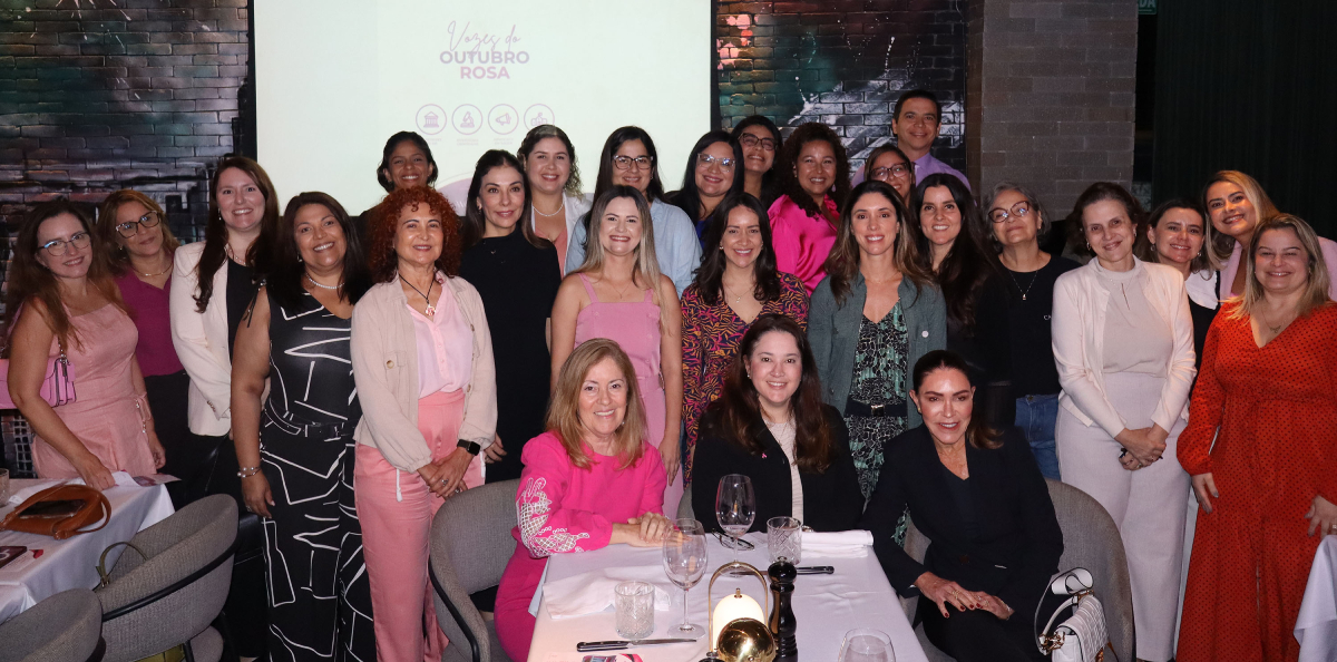 Fotografia de evento com muitas pessoas adultas sorrindo diante de tela com o texto “Viva o Outubro Rosa”, reunidas em ambiente interno iluminado.