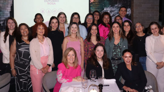 Fotografia de evento com muitas pessoas adultas sorrindo diante de tela com o texto “Viva o Outubro Rosa”, reunidas em ambiente interno iluminado.