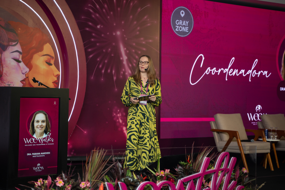 Coordenadora apresenta conteúdo no palco diante de uma tela com sua identificação e o cenário do WEEM Talks 2026.
