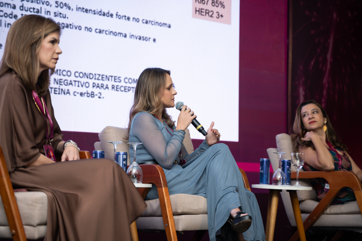 Três participantes sentadas no palco durante uma discussão de painel, com uma delas falando ao microfone.