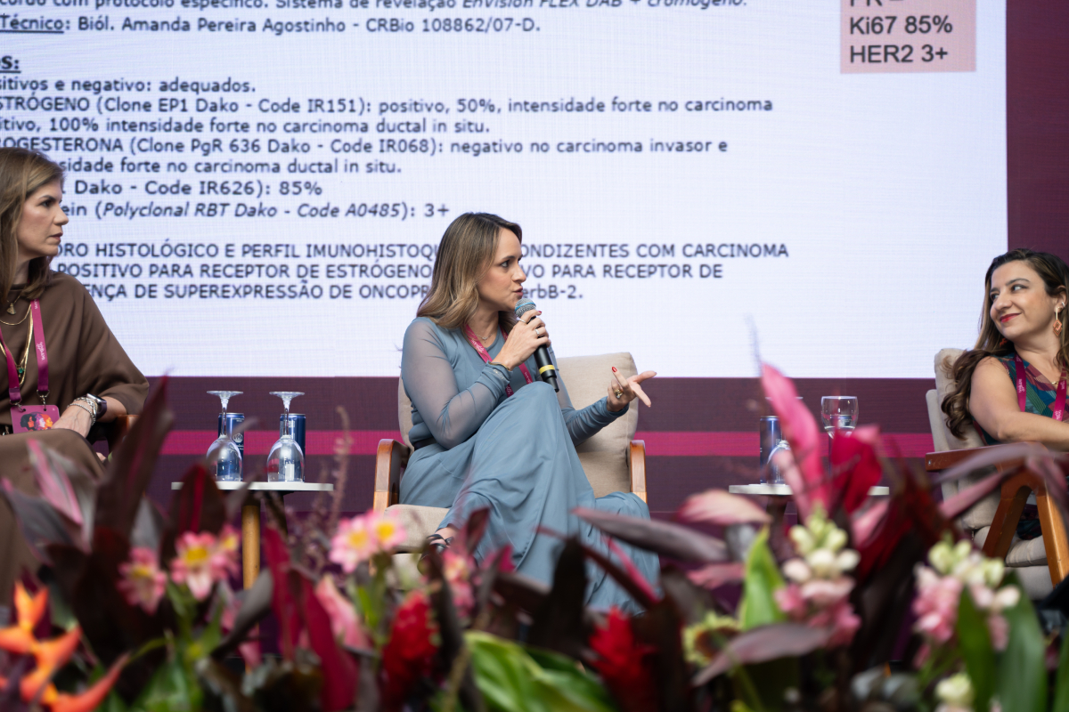 Participante fala ao microfone durante painel, com conteúdo médico exibido em uma tela ao fundo.