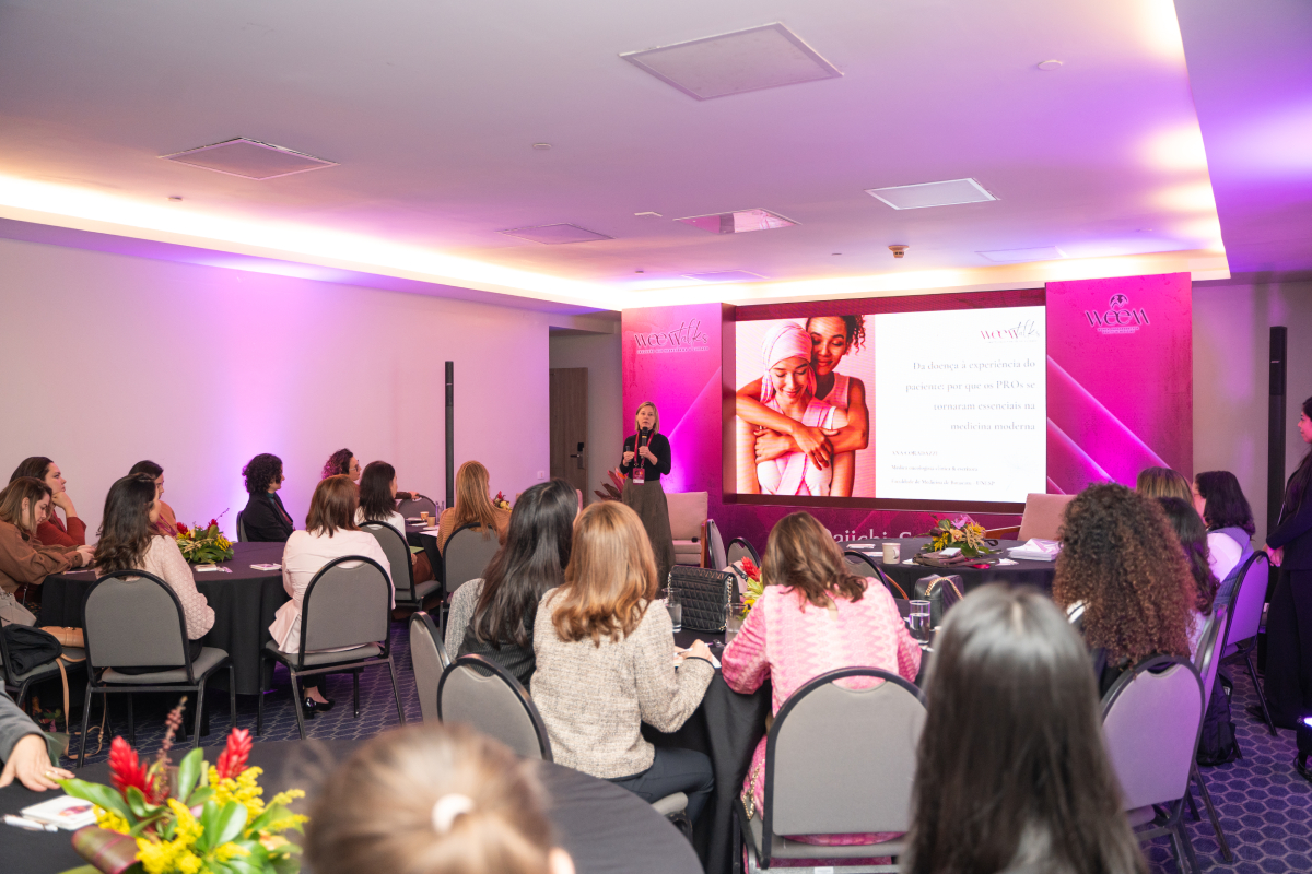 Palestrante apresenta conteúdo sobre jornada e participação de pacientes, enquanto a plateia acompanha em uma sala do WEEM Talks 2026.