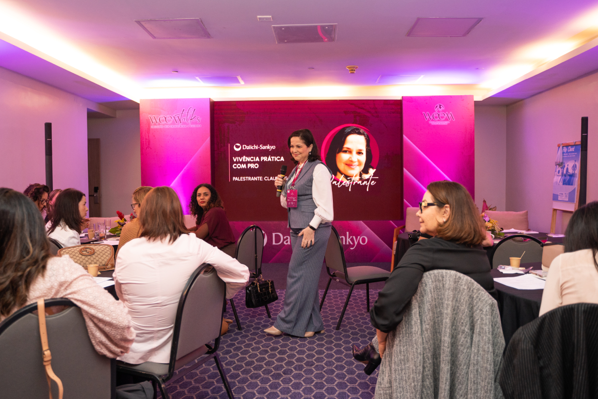 Palestrante conduz uma apresentação em sala com participantes sentadas em mesas, diante de uma tela com conteúdo institucional.