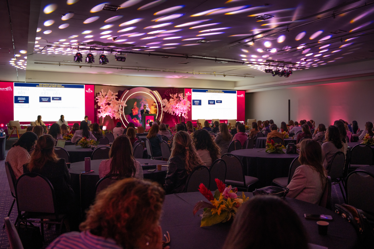 Vista ampla do auditório com participantes sentados em mesas e o palco do WEEM Talks 2026 ao fundo.