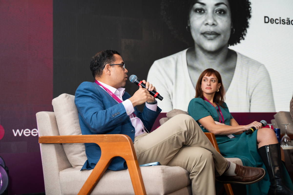Participante fala ao microfone durante painel, enquanto outra participante acompanha a discussão sentada ao lado.
